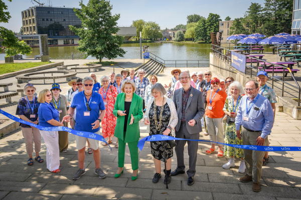 Diana Moran cutting the ribbon at u3a Festival 24. Behind her, there is a crowd of u3a members and the lake at the University of York.